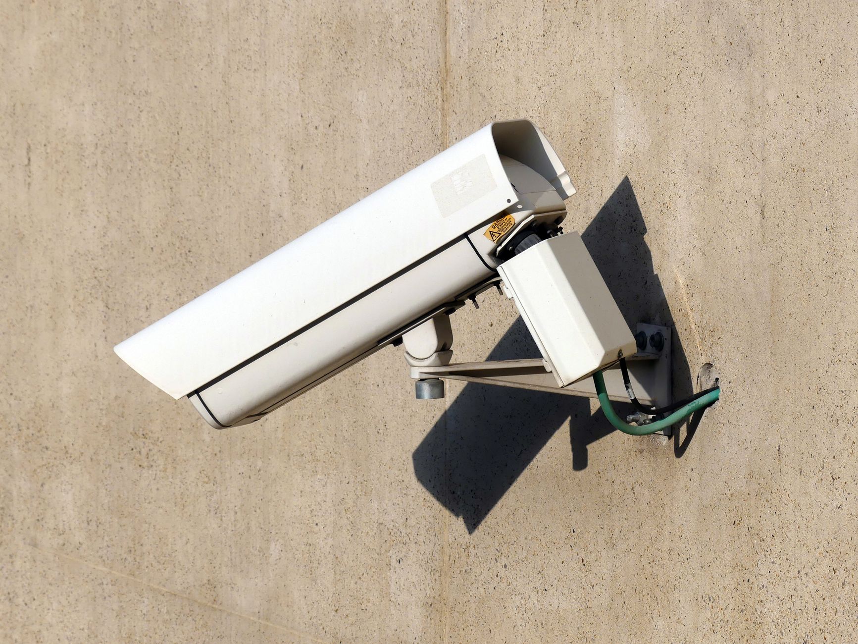 Close-up of a white security camera mounted on a beige wall in sunlight.