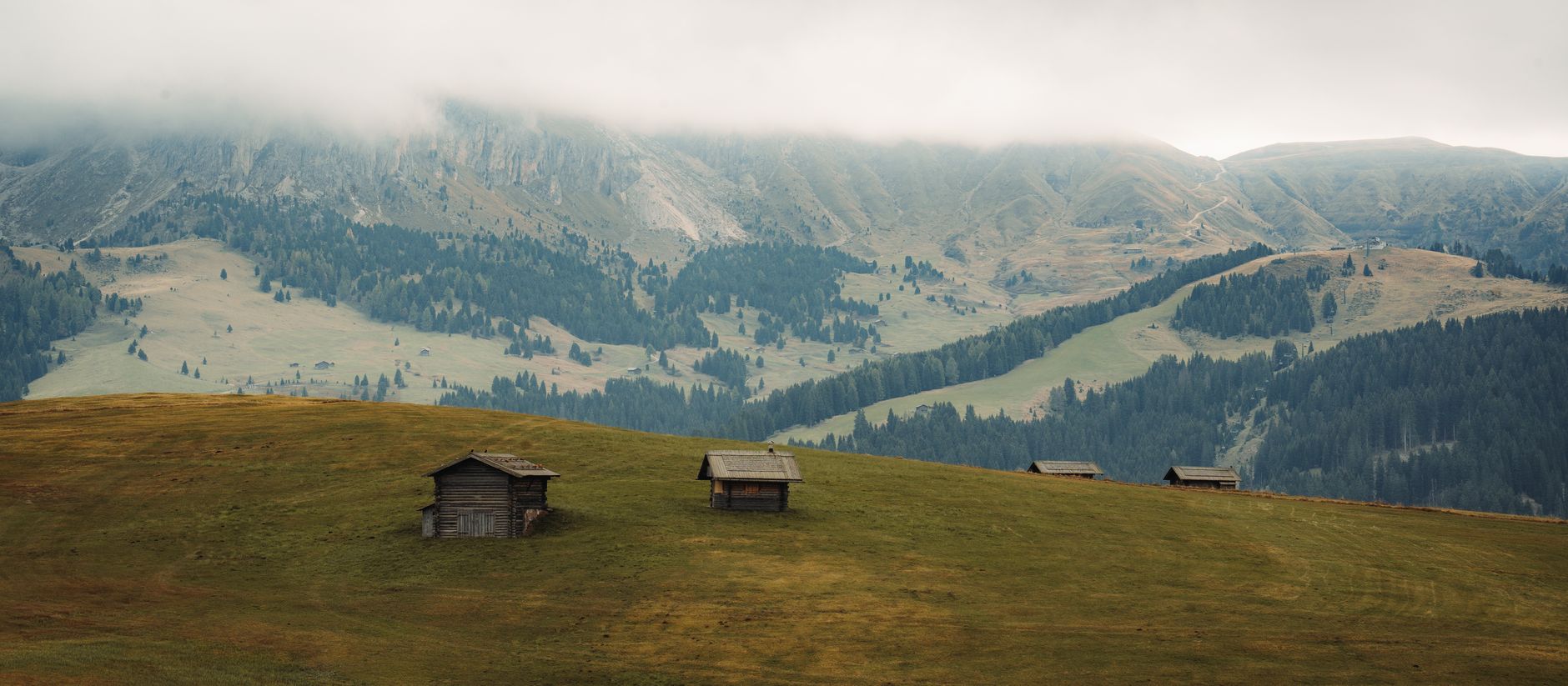 Wooden cabins on a misty alpine meadow with forested mountains